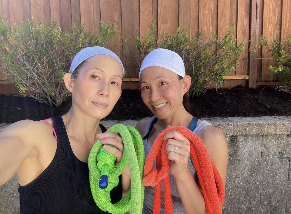 Twin sisters Sandy and Candy Fong outdoors in workout tanks and headbands, each holding a coiled jump rope, one green and one orange.