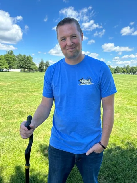 Jay Armstrong smiling in a blue T-shirt, holding a black cane on a sunny grassy field.