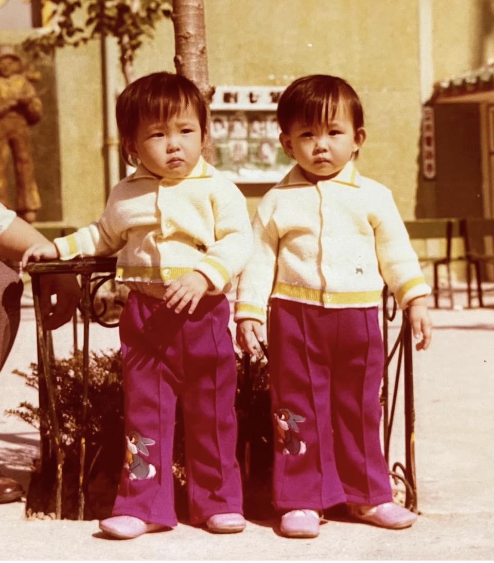 Vintage color photo of twin toddler girls in matching cardigans and patterned pants, standing side by side outdoors.