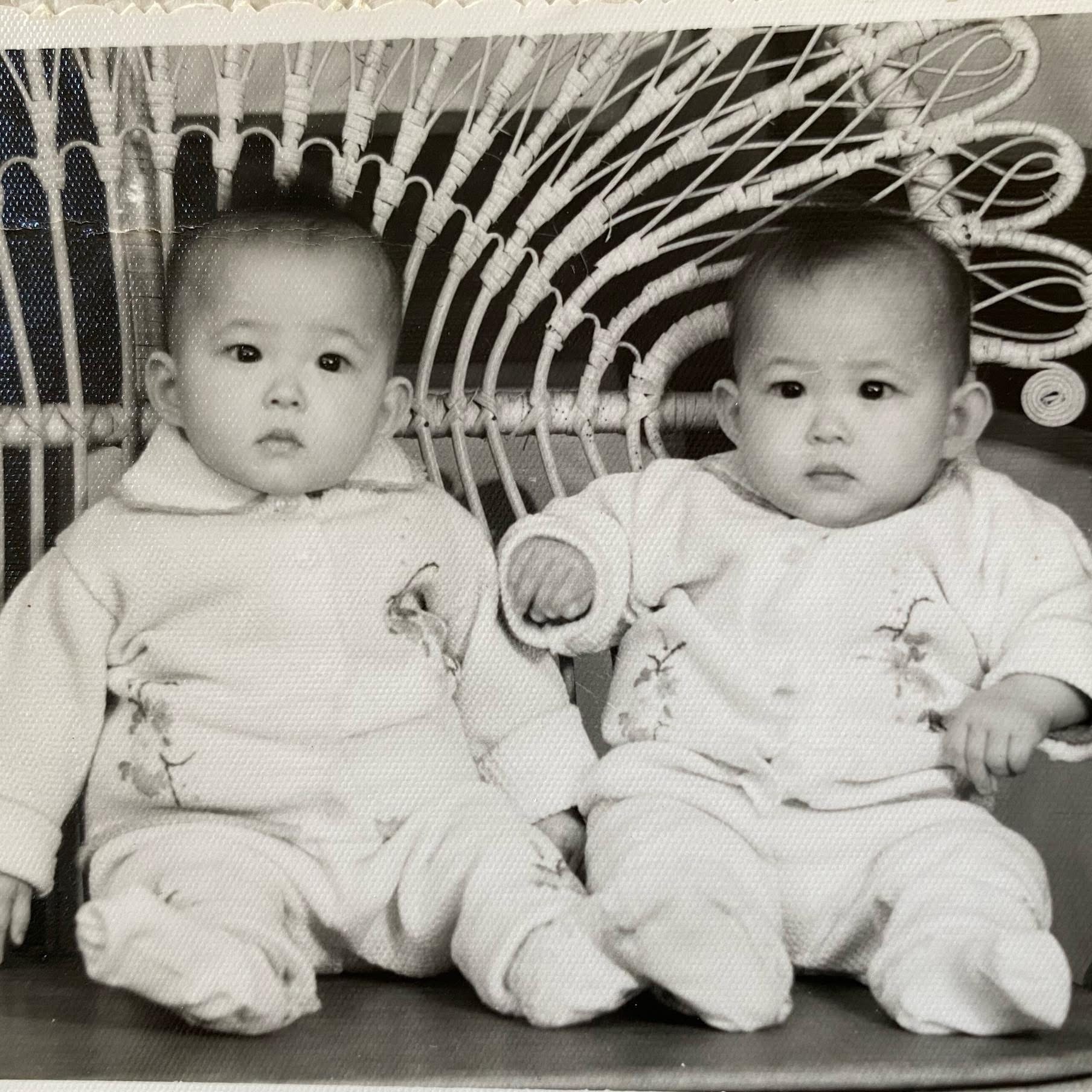 Black and white photo of twin baby girls in matching white knit outfits, seated side by side in a wicker chair.