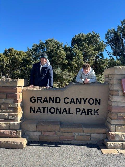 Jay and his son leaning over the Grand Canyon National Park entrance sign.